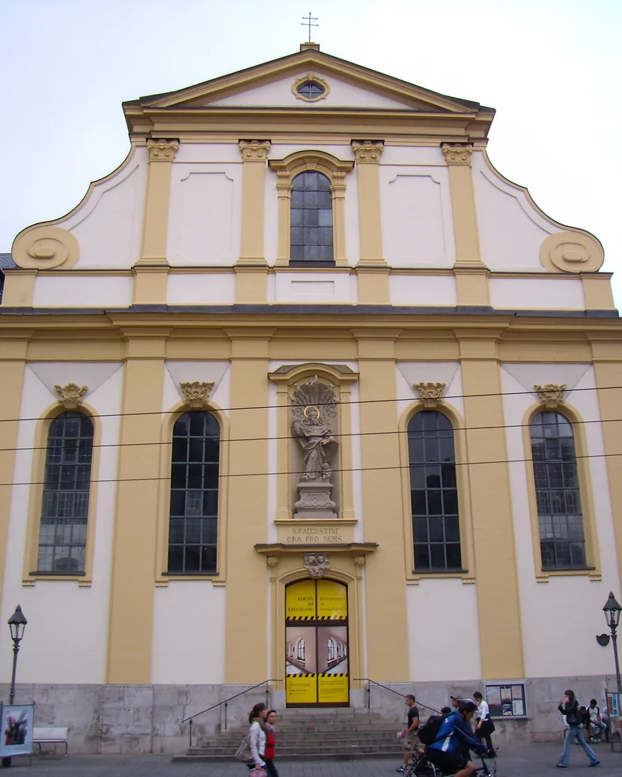 Die Augustinerkirche am Dominikanerplatz, gelb-weiße Fassade mit geschwungenem Giebel und Heiligenfigur über dem Eingang