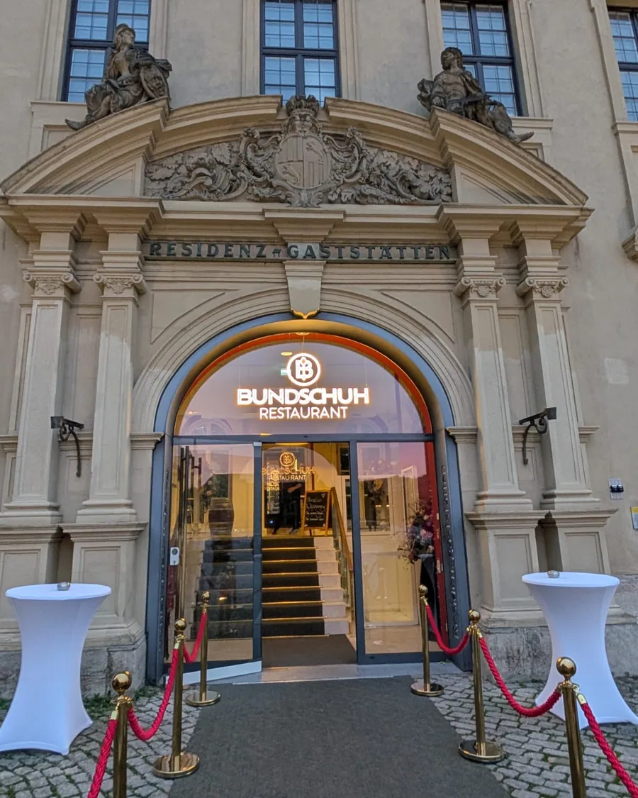 Das barocke Portal der Residenzgaststätten mit der Leuchtschrift Bundschuh Restaurant über der Glastür, davor ein Teppich zwischen roten Kordeln