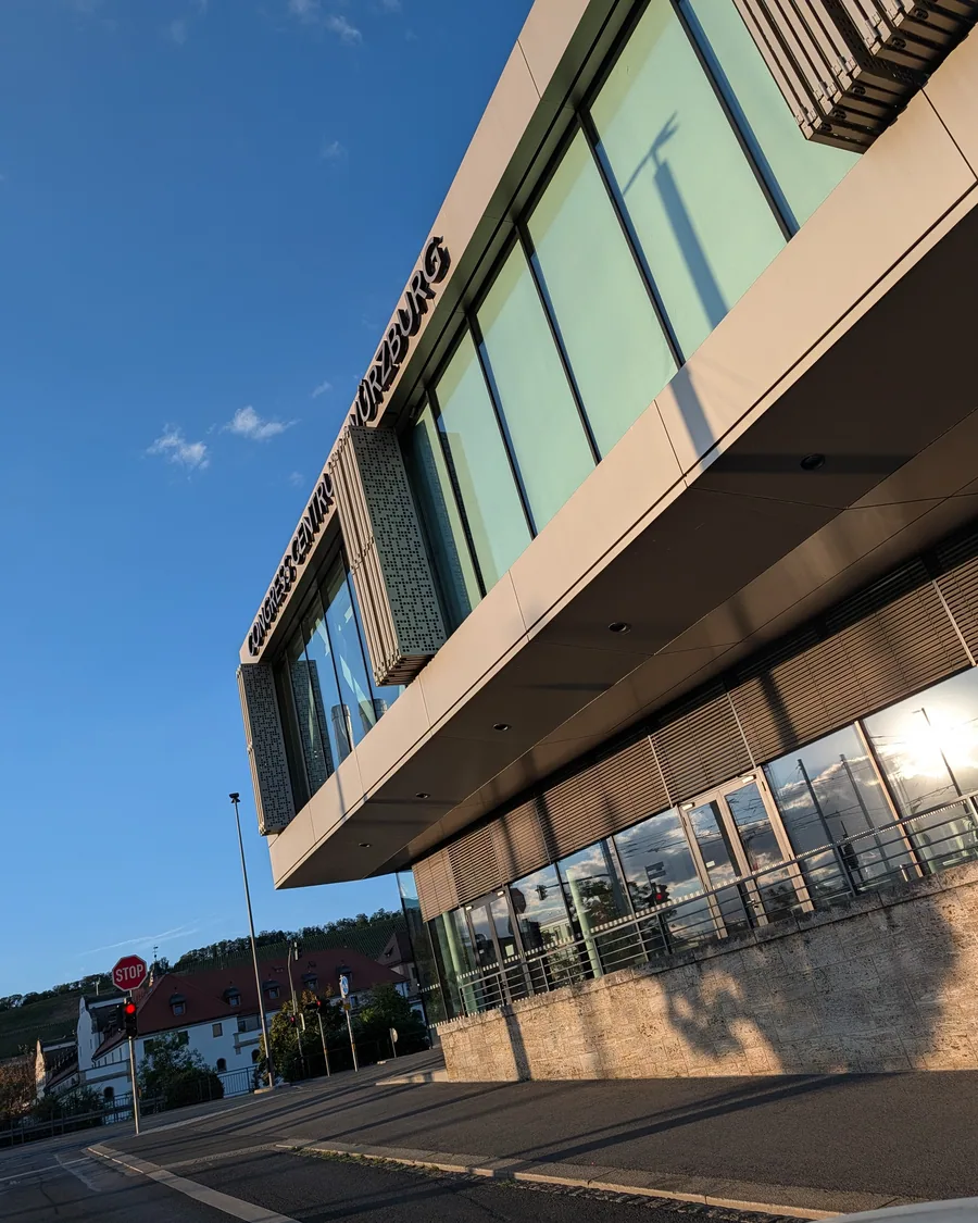Die auskragende Glasfassade des Congress Centrums Würzburg von unten, mit dem Schriftzug am Dachrand, dahinter Weinberge im Abendlicht