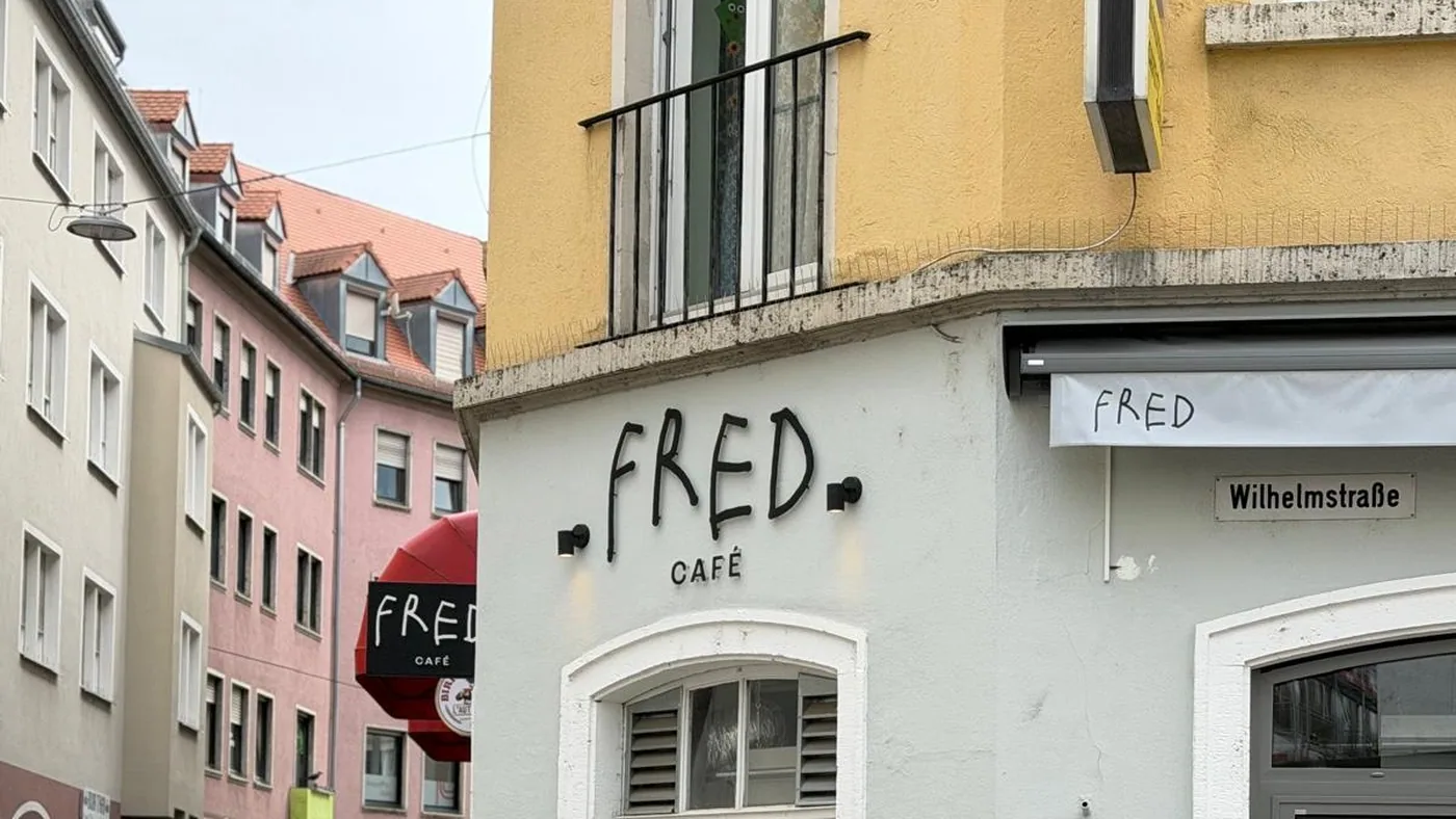 Der handgemalte Schriftzug FRED CAFÉ an der hellen Eckfassade in der Herzogenstraße, daneben das Straßenschild Wilhelmstraße und zwei Tafeln mit den Angeboten des Tages
