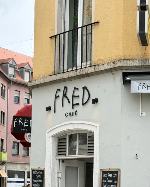 Der handgemalte Schriftzug FRED CAFÉ an der hellen Eckfassade in der Herzogenstraße, daneben das Straßenschild Wilhelmstraße und zwei Tafeln mit den Angeboten des Tages