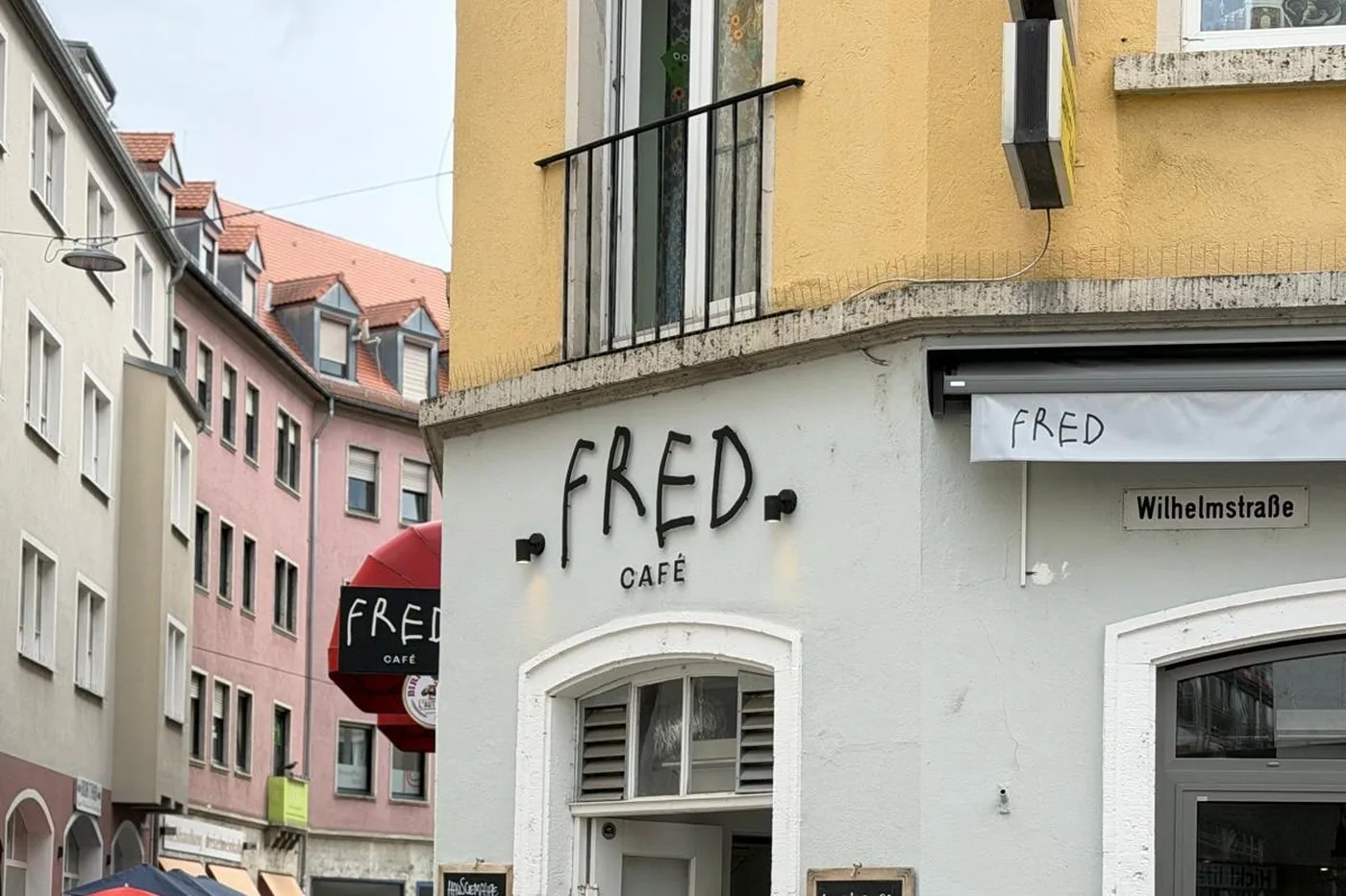 Der handgemalte Schriftzug FRED CAFÉ an der hellen Eckfassade in der Herzogenstraße, daneben das Straßenschild Wilhelmstraße und zwei Tafeln mit den Angeboten des Tages
