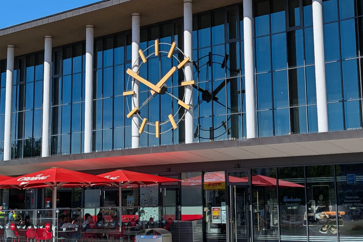 Die Bahnhofsuhr am Wuerzburger Hauptbahnhof aus der Naehe, davor die roten Sonnenschirme des Bahnhofscafes