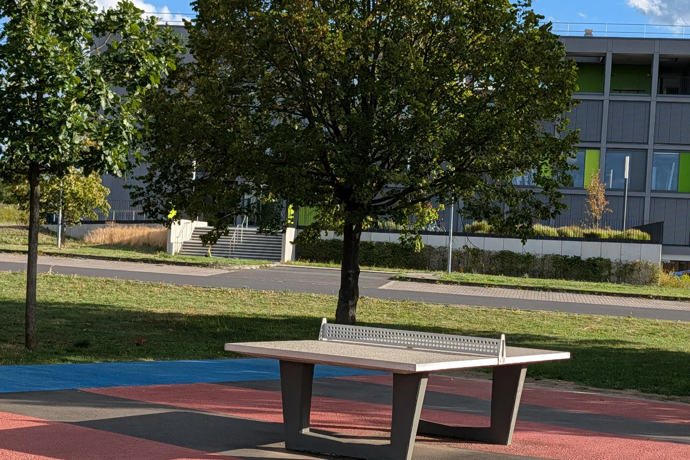 Eine Tischtennisplatte aus Beton auf rot und grau gestreiftem Belag am Hubland, dahinter ein Baum und ein Schulgebäude