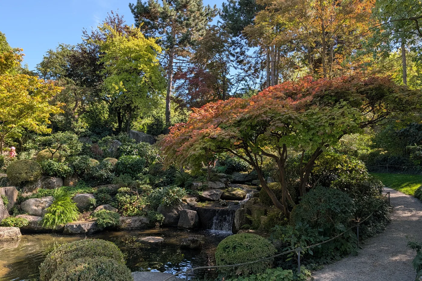 Eine hohe Steinlaterne am Teich, dahinter der Wasserfall zwischen Findlingen und geschnittenen Sträuchern
