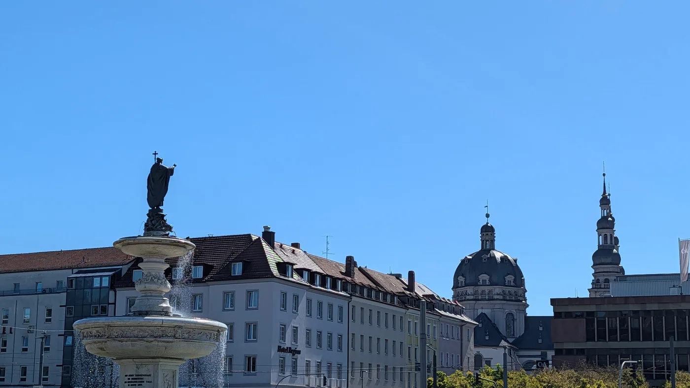 Der Kiliansbrunnen vor dem Hauptbahnhof, dahinter die Kuppel des Stift Haug und die Daecher der Innenstadt