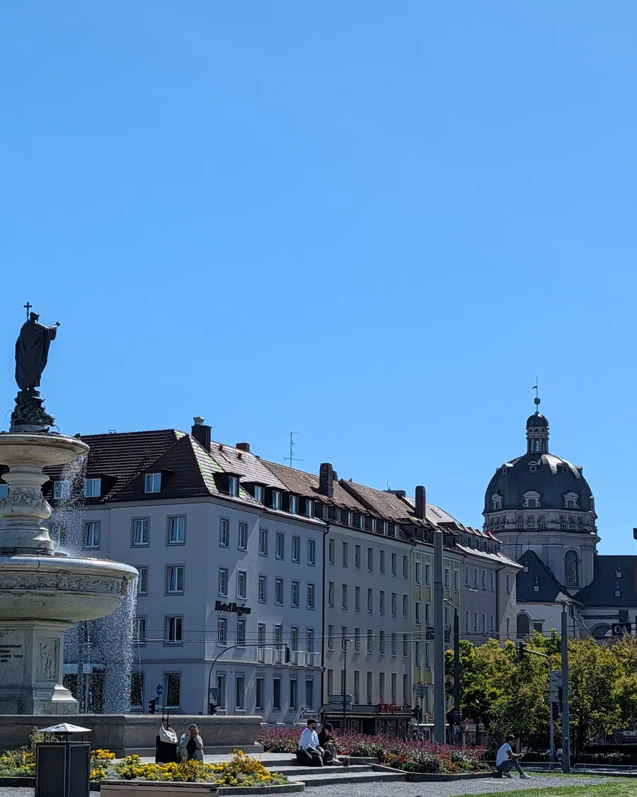 Der Kiliansbrunnen vor dem Hauptbahnhof, dahinter die Kuppel des Stift Haug und die Daecher der Innenstadt