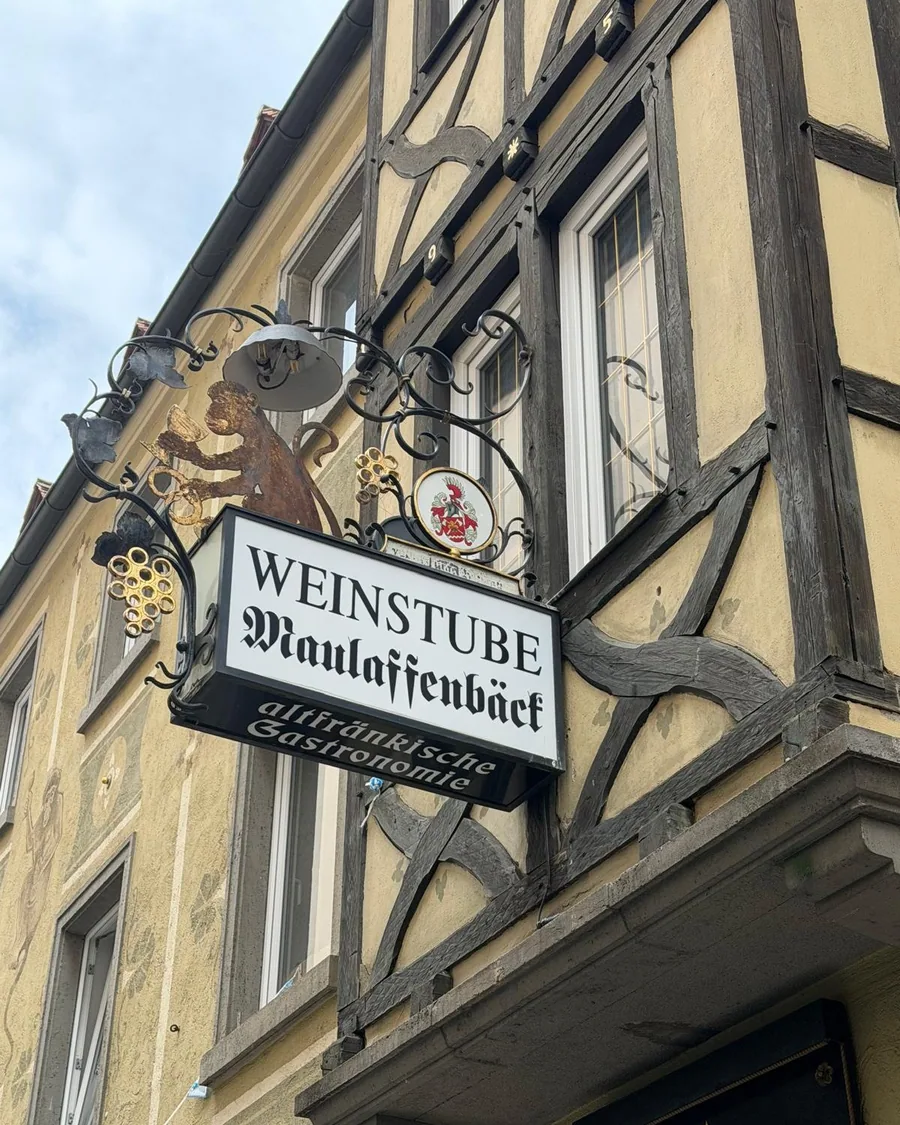 Das schmiedeeiserne Wirtshausschild der Weinstube Maulaffenbäck mit dem lesenden Affen und dem Würzburger Wappen, am Fachwerk der Maulhardgasse