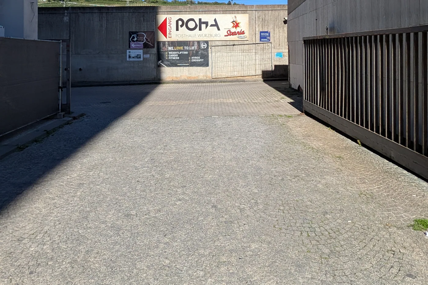 Die Zufahrt zur Posthalle zwischen den Hallen, am Ende der Wegweiser zum Eingang, dahinter die Bahnstrecke und die Weinberge