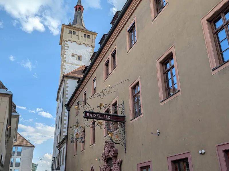 Blick die Gasse hinauf zum Eingang des Ratskellers im Würzburger Rathaus, mit dem Nasenschild über der Tür und dem Rathausturm dahinter