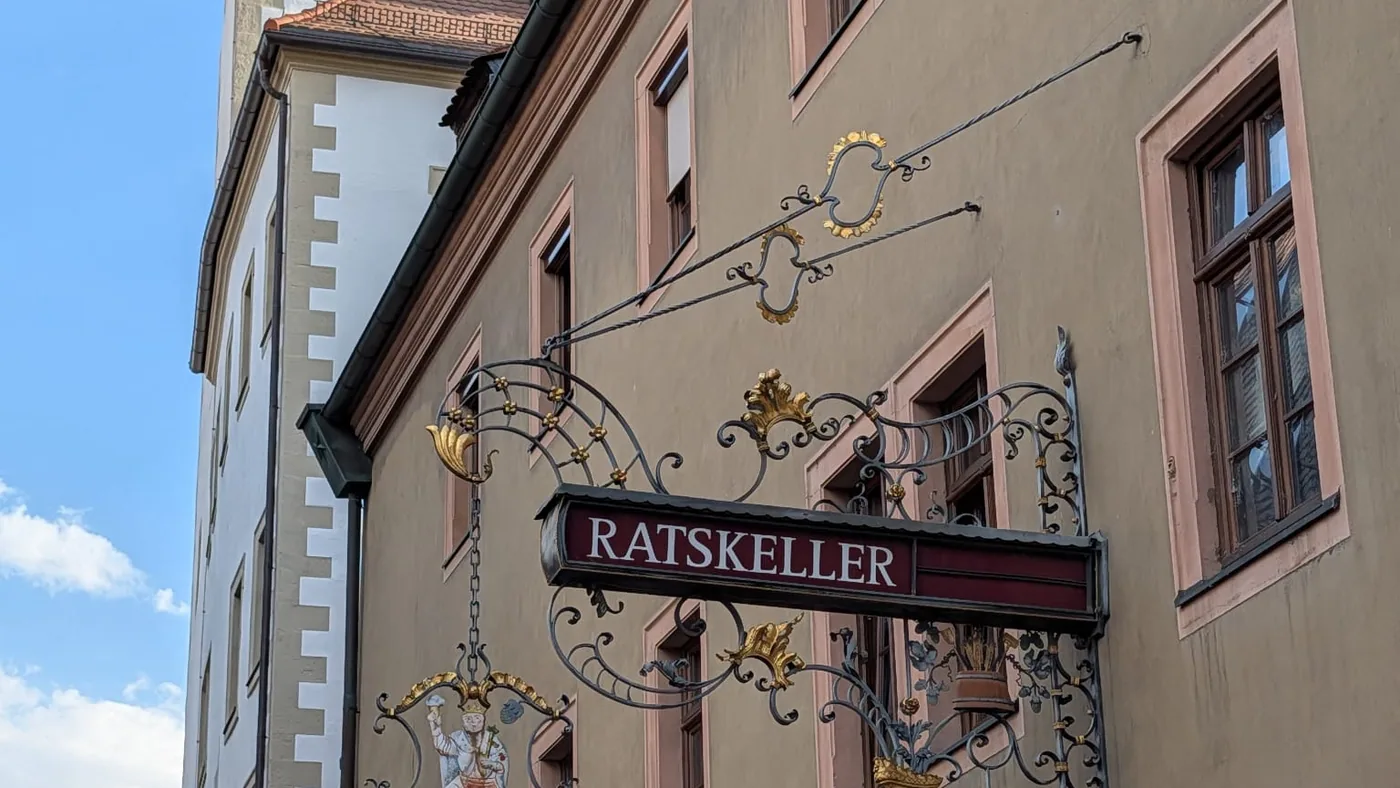 Das schmiedeeiserne Nasenschild des Ratskellers mit einer Bacchusfigur auf einem Weinfass und dem Würzburger Wappen, dahinter der Turm des Rathauses