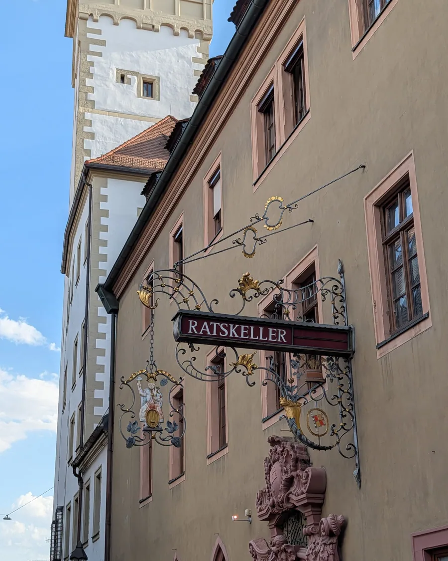 Das schmiedeeiserne Nasenschild des Ratskellers mit einer Bacchusfigur auf einem Weinfass und dem Würzburger Wappen, dahinter der Turm des Rathauses