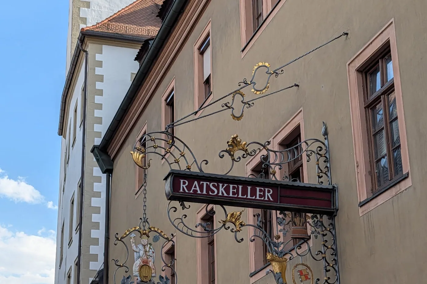 Das schmiedeeiserne Nasenschild des Ratskellers mit einer Bacchusfigur auf einem Weinfass und dem Würzburger Wappen, dahinter der Turm des Rathauses