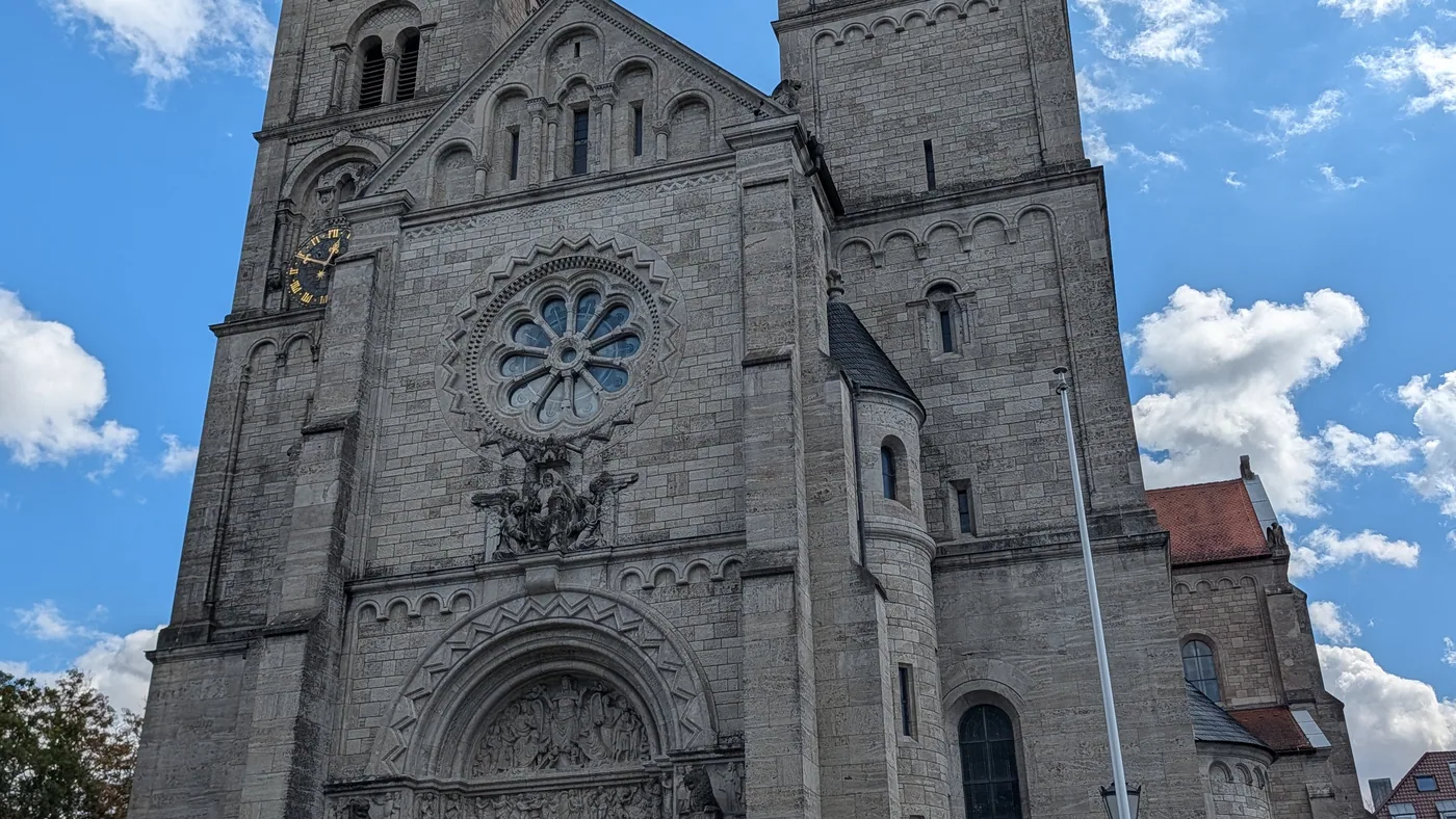 Die Westfassade von St. Adalbero in der Würzburger Sanderau, zwei hohe Türme mit spitzen Helmen und eine große Fensterrose über dem Stufenportal