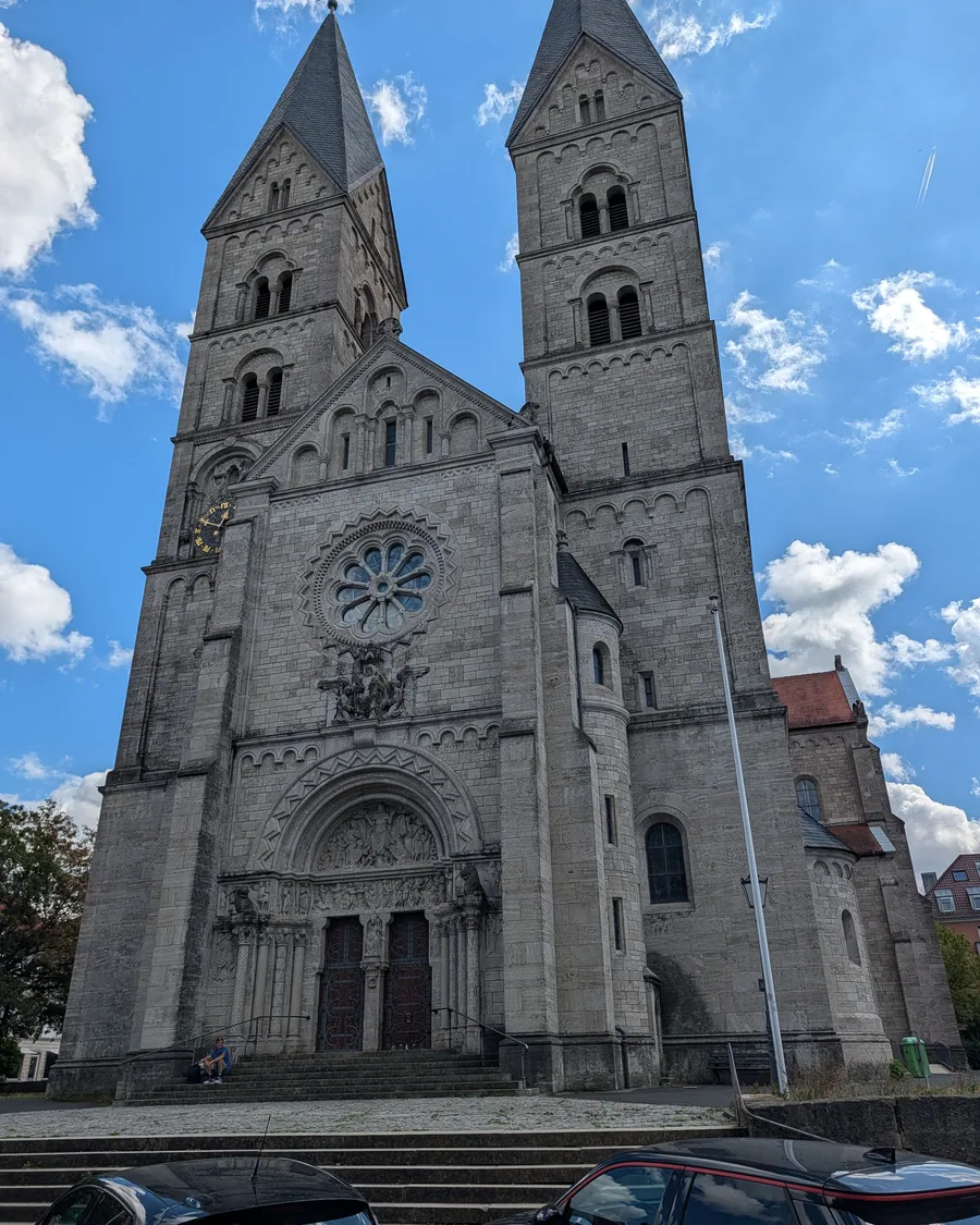 Die Westfassade von St. Adalbero in der Würzburger Sanderau, zwei hohe Türme mit spitzen Helmen und eine große Fensterrose über dem Stufenportal