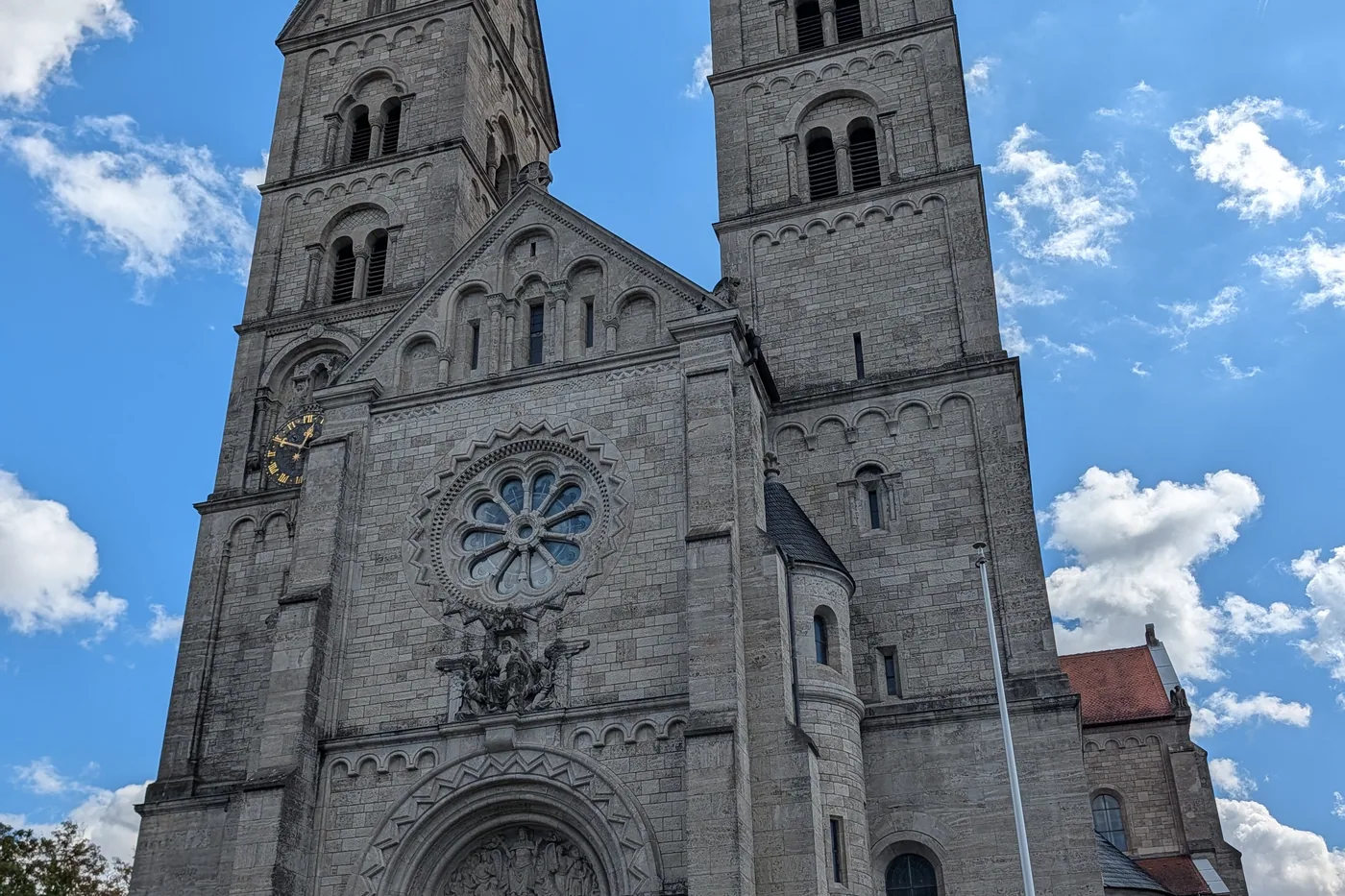 Die Westfassade von St. Adalbero in der Würzburger Sanderau, zwei hohe Türme mit spitzen Helmen und eine große Fensterrose über dem Stufenportal