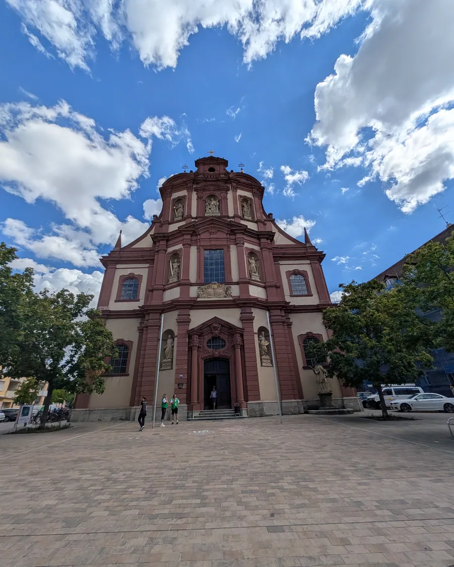 Die barocke Fassade der Kirche St. Peter und Paul in Würzburg, rot und weiß gefasst, mit Heiligenfiguren in Nischen über dem Portal