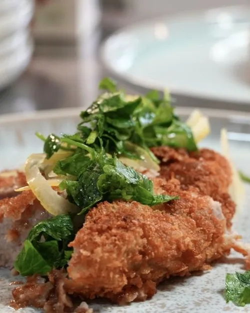 Gebackenes Fleisch mit Kräutersalat auf einem hellen Teller, im Hintergrund ein zweiter Teller und ein Stapel Geschirr