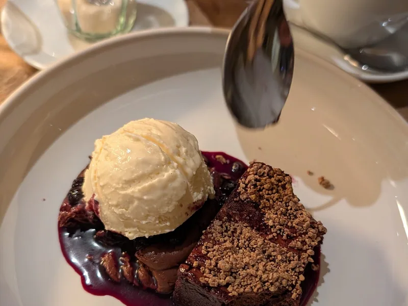 Ein Stück dunkler Kuchen mit Streuseln neben einer Kugel hellem Eis auf dunkelrotem Fruchtspiegel, dahinter eine Tasse Kaffee mit Milchschaum
