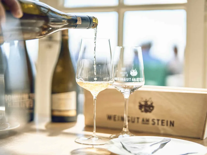 Weißwein wird in ein Glas mit dem Schriftzug Weingut am Stein eingeschenkt, daneben eine Holzkiste des Weinguts