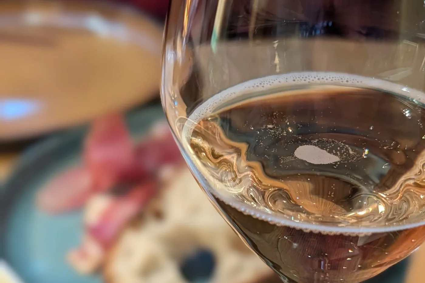 Ein Glas mit hellem Wein im Vordergrund, dahinter unscharf ein Teller mit geröstetem Brot und dünn aufgelegtem Fleisch