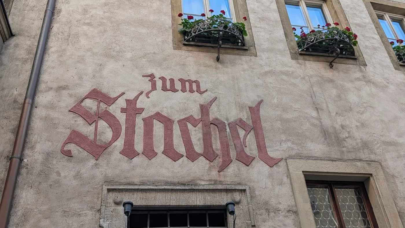 Der Schriftzug Zum Stachel in roter Fraktur an der Fassade des Weinhauses, darüber Fenster mit roten Geranien